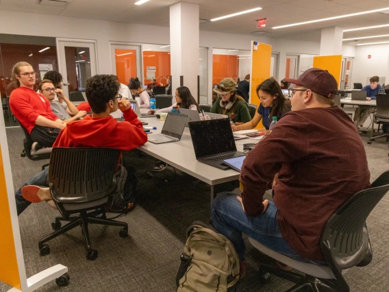 students sitting at a table writing, talking, working on laptops